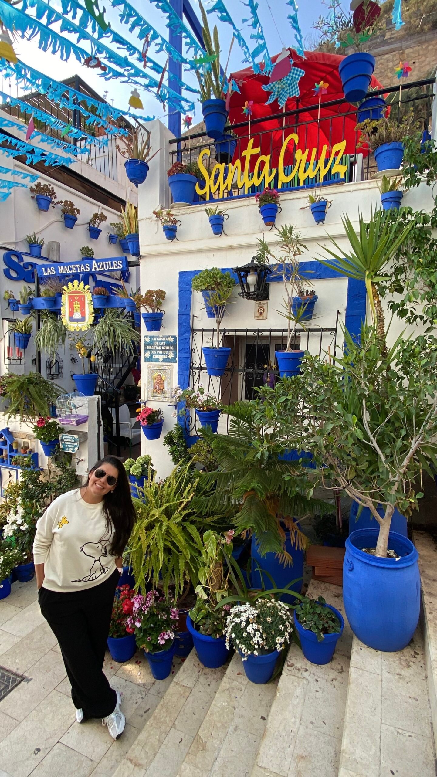 posando sonriente en las escaleras del pintoresco Rinc&oacute;n de las Macetas Azules en el Barrio de Santa Cruz, Alicante. La fachada blanca de la casa est&aacute; decorada con maceteros azules colgados en la pared, un letrero amarillo que dice "Santa Cruz" y plantas verdes t&iacute;picas del casco antiguo