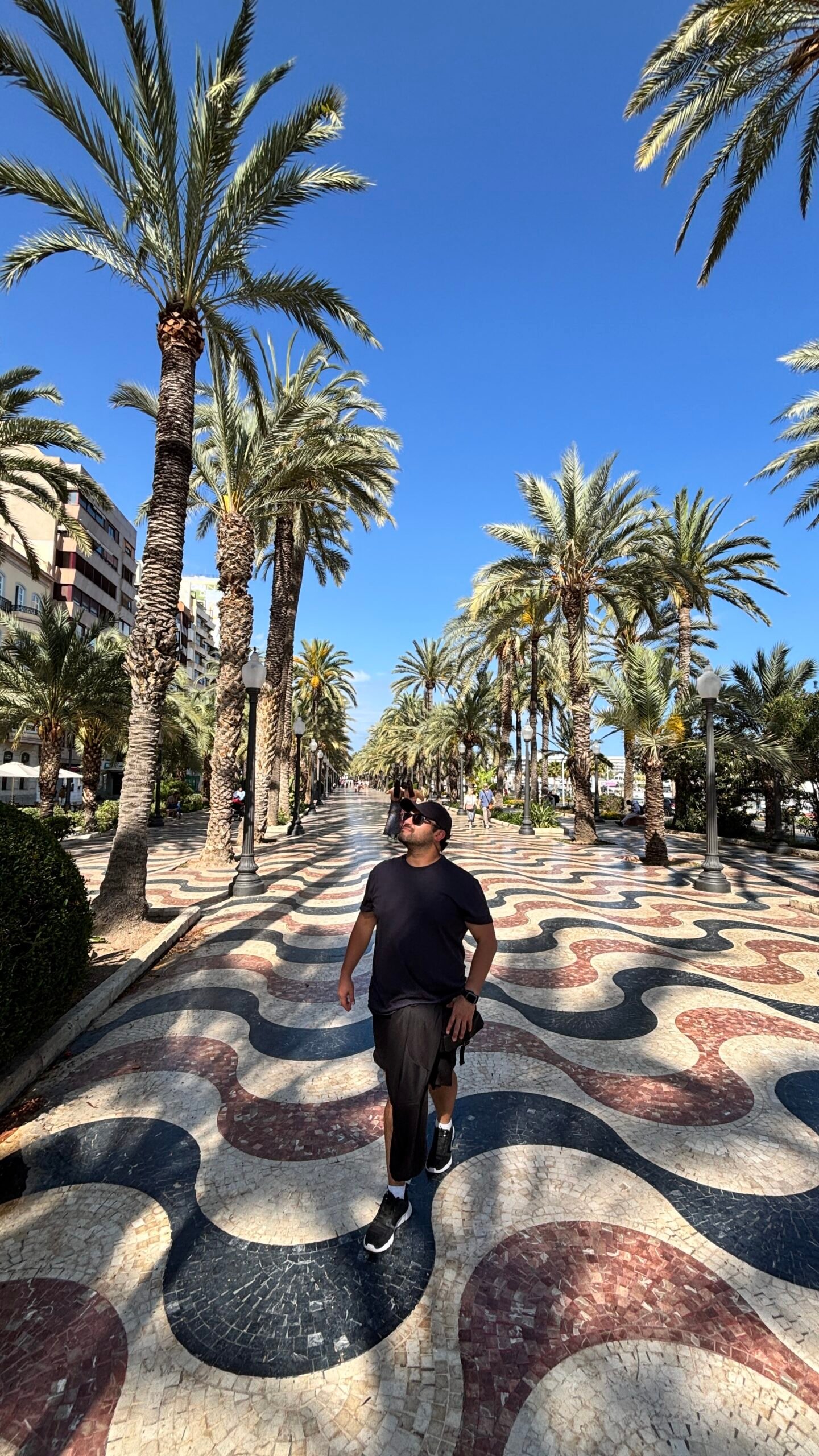 caminando y mirando hacia arriba en el famoso paseo de la Explanada de Espa&ntilde;a en Alicante. Viste ropa deportiva negra con gorra y gafas de sol. De fondo se aprecia el caracter&iacute;stico suelo de mosaico ondulado en tonos rojo, azul y blanco, flanqueado por hileras de altas palmeras y farolas cl&aacute;sicas bajo un d&iacute;a soleado.