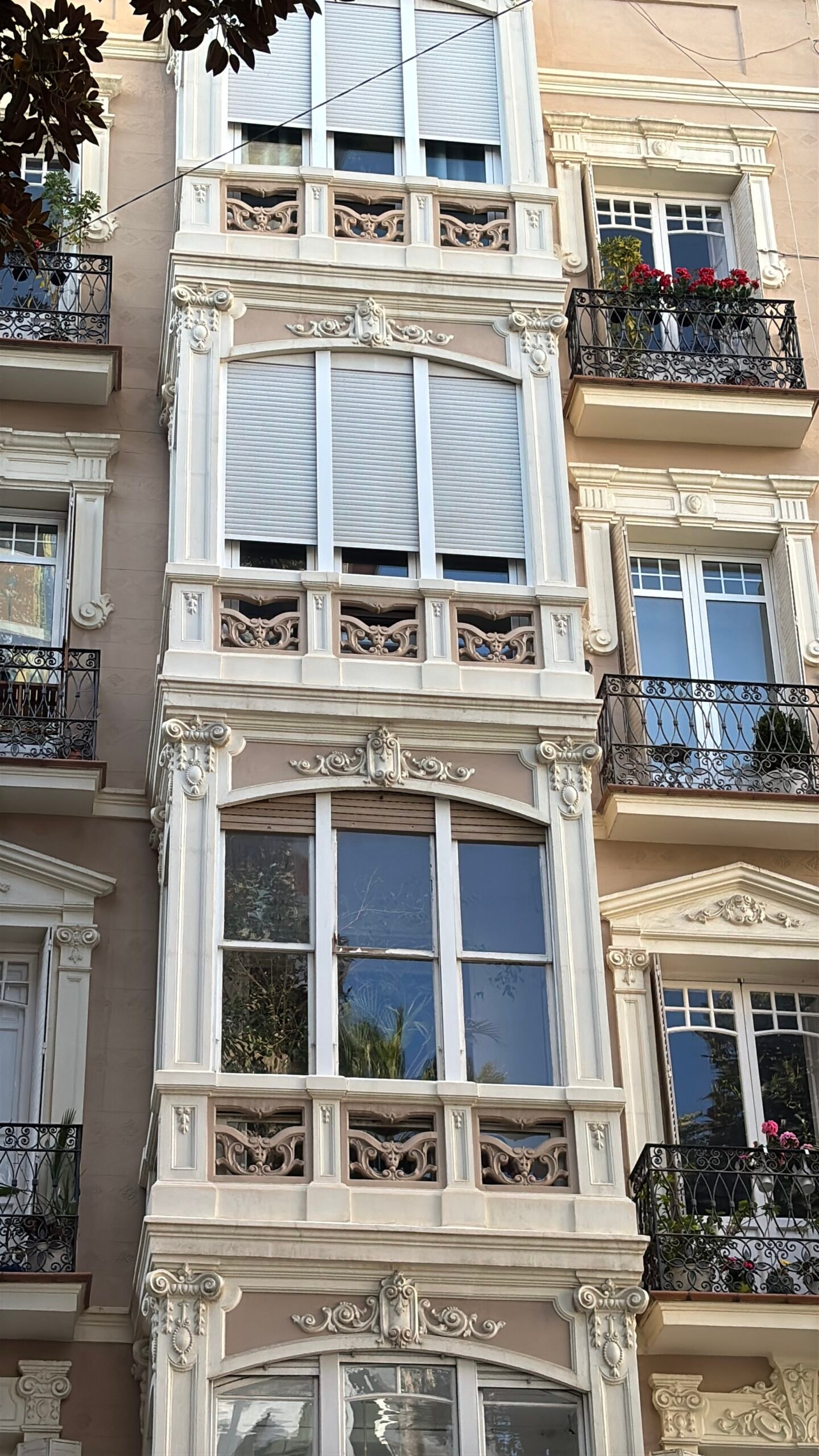Primer plano de una fachada de edificio modernista en Alicante. La imagen captura los intrincados detalles arquitect&oacute;nicos de color blanco y beige, incluyendo balcones de hierro forjado con flores rojas, ventanas ornamentadas y elementos decorativos esculpidos alrededor de los ventanales. El estilo es cl&aacute;sico de principios del siglo XX.