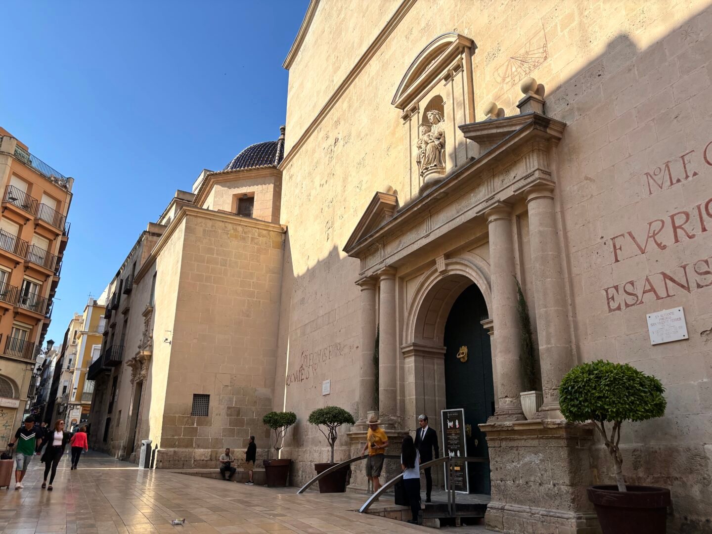 Vista exterior de la imponente fachada de piedra de la Concatedral de San Nicol&aacute;s de Bari en el centro hist&oacute;rico de Alicante. Muestra la portada principal de estilo renacentista, la c&uacute;pula de azulejos azules al fondo y la plaza peatonal con transe&uacute;ntes bajo un cielo azul despejado.