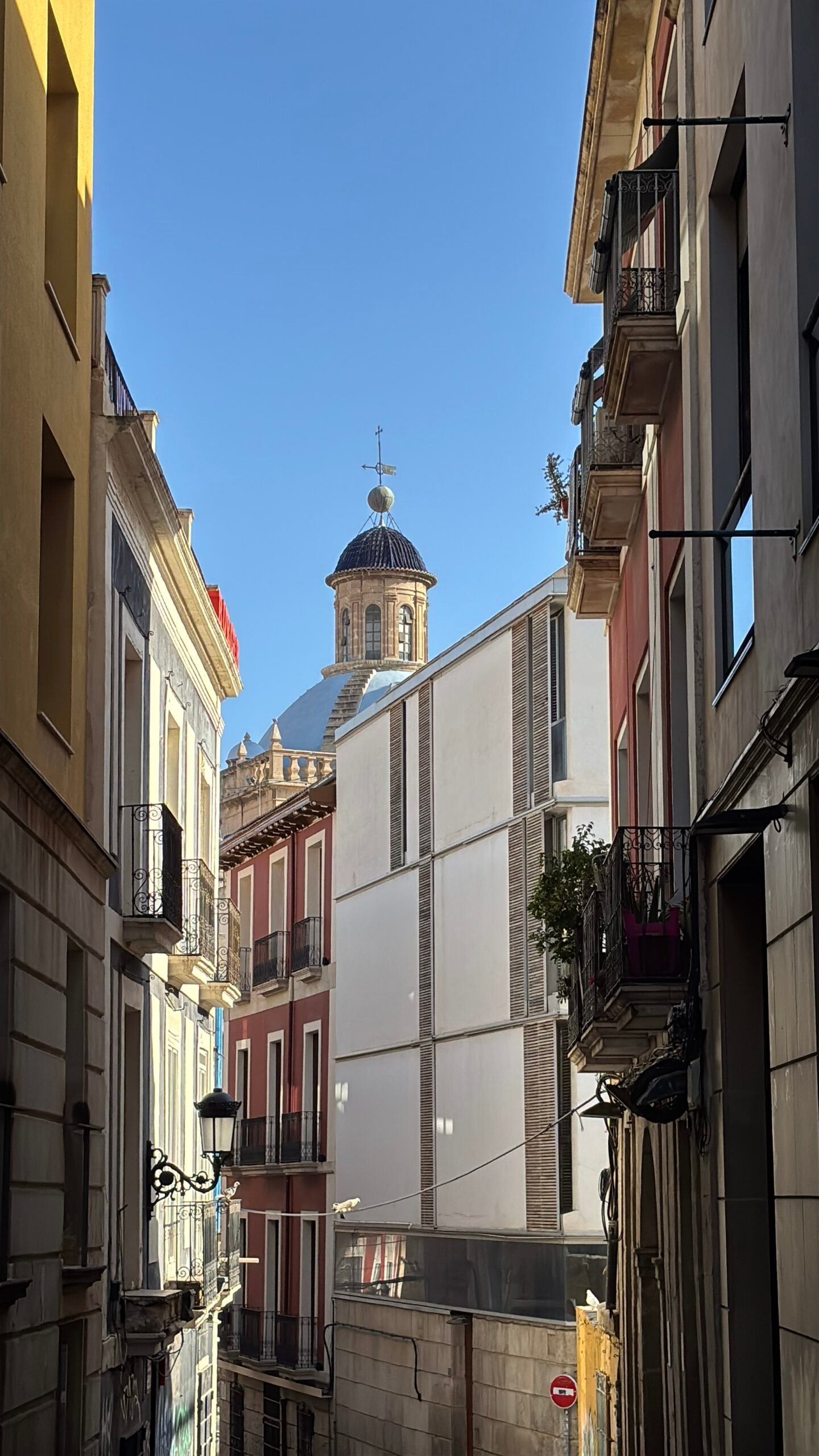 Una toma en &aacute;ngulo de un callej&oacute;n estrecho en el casco antiguo de Alicante. Las paredes de los edificios tradicionales de diferentes colores enmarcan la vista. Al fondo, bajo un cielo azul despejado, se alza la c&uacute;pula de azulejos azules y beige de una iglesia hist&oacute;rica (posiblemente la Concatedral de San Nicol&aacute;s).