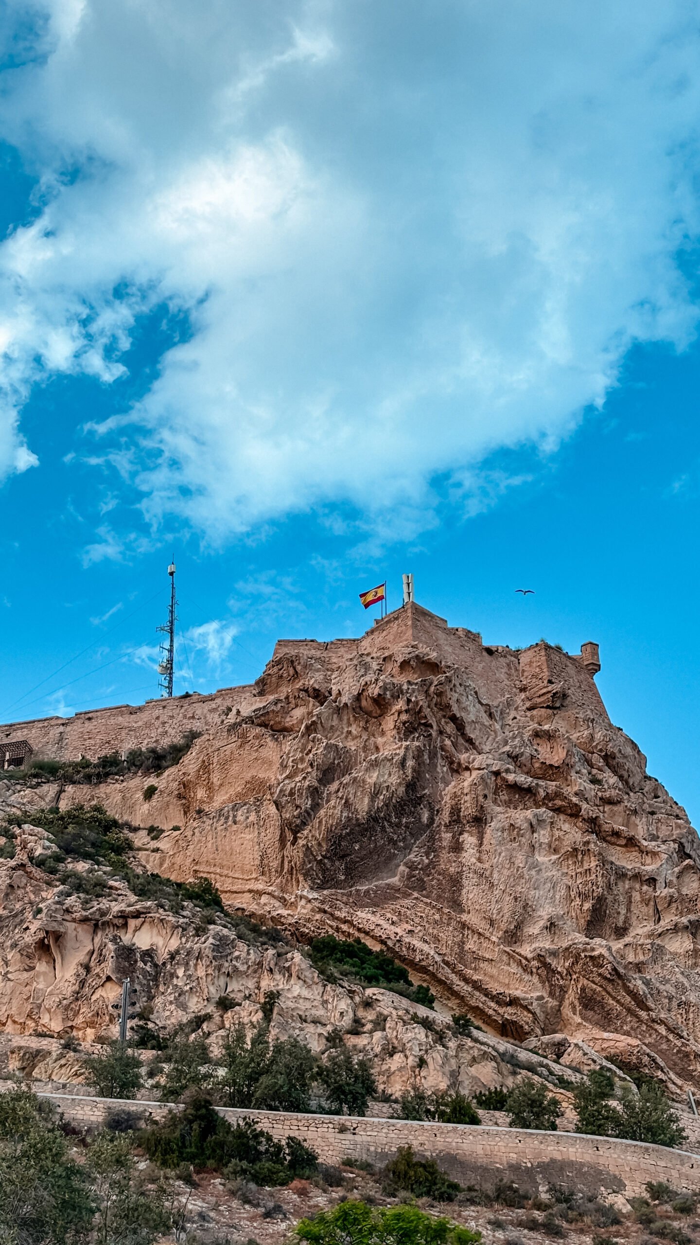 Vista panor&aacute;mica y elevada de las murallas e historia del Castillo de Santa B&aacute;rbara, situado en la cima del imponente acantilado rocoso del Monte Benacantil en Alicante, Espa&ntilde;a. La bandera de Espa&ntilde;a ondea en la cumbre de la fortaleza militar bajo un cielo azul con nubes blancas.