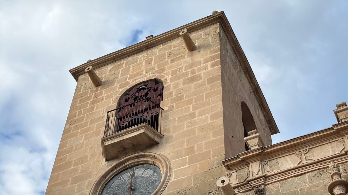 Primer plano de la torre del campanario de piedra de la Bas&iacute;lica de Santa Mar&iacute;a en Alicante, mostrando un reloj circular en la parte inferior, un ventanal con balc&oacute;n de hierro forjado y un cielo ligeramente nublado de fondo.