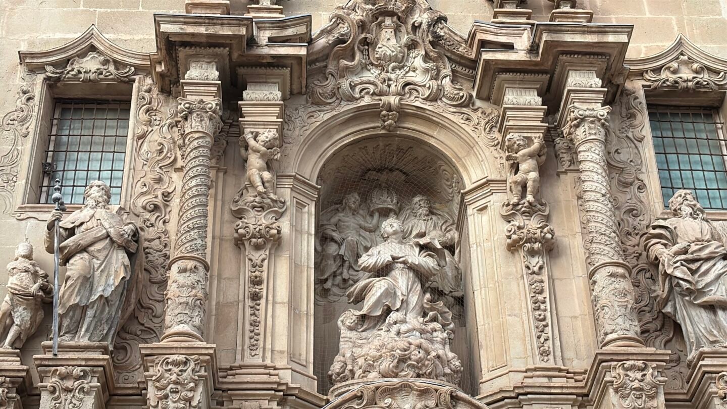 Detalle en primer plano de la fachada principal barroca de la Bas&iacute;lica de Santa Mar&iacute;a en Alicante. Muestra intrincadas tallas en piedra con estatuas de santos, hornacinas ornamentadas y columnas esculpidas de estilo barroco espa&ntilde;ol.