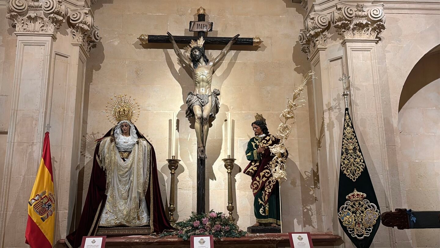 Capilla interior de la Bas&iacute;lica de Santa Mar&iacute;a en Alicante con un altar que alberga una escultura de Cristo Crucificado en el centro, flanqueado por la Virgen Mar&iacute;a vestida de luto y la estatua de un santo, junto a la bandera de Espa&ntilde;a y un estandarte procesional.