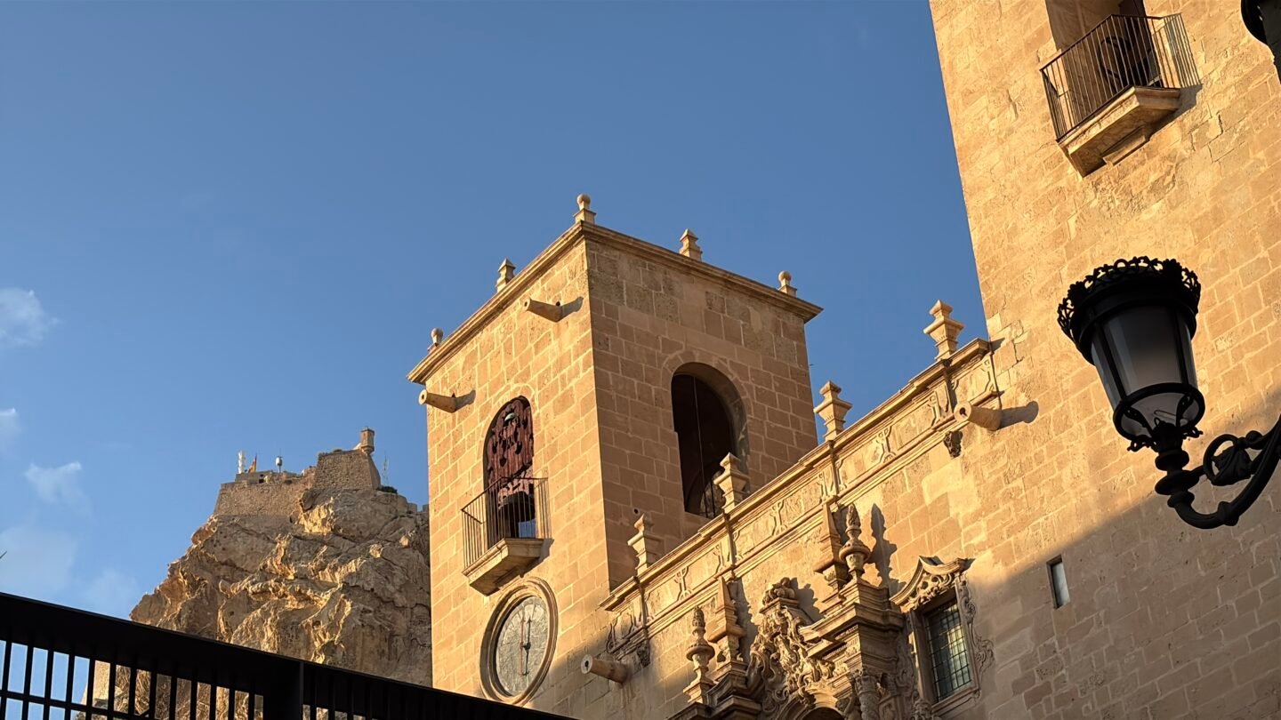 Vista en perspectiva de la fachada de piedra dorada y la torre de la Bas&iacute;lica de Santa Mar&iacute;a en Alicante iluminada por el sol, con la impresionante cumbre rocosa del Castillo de Santa B&aacute;rbara visible al fondo bajo un cielo azul despejado.