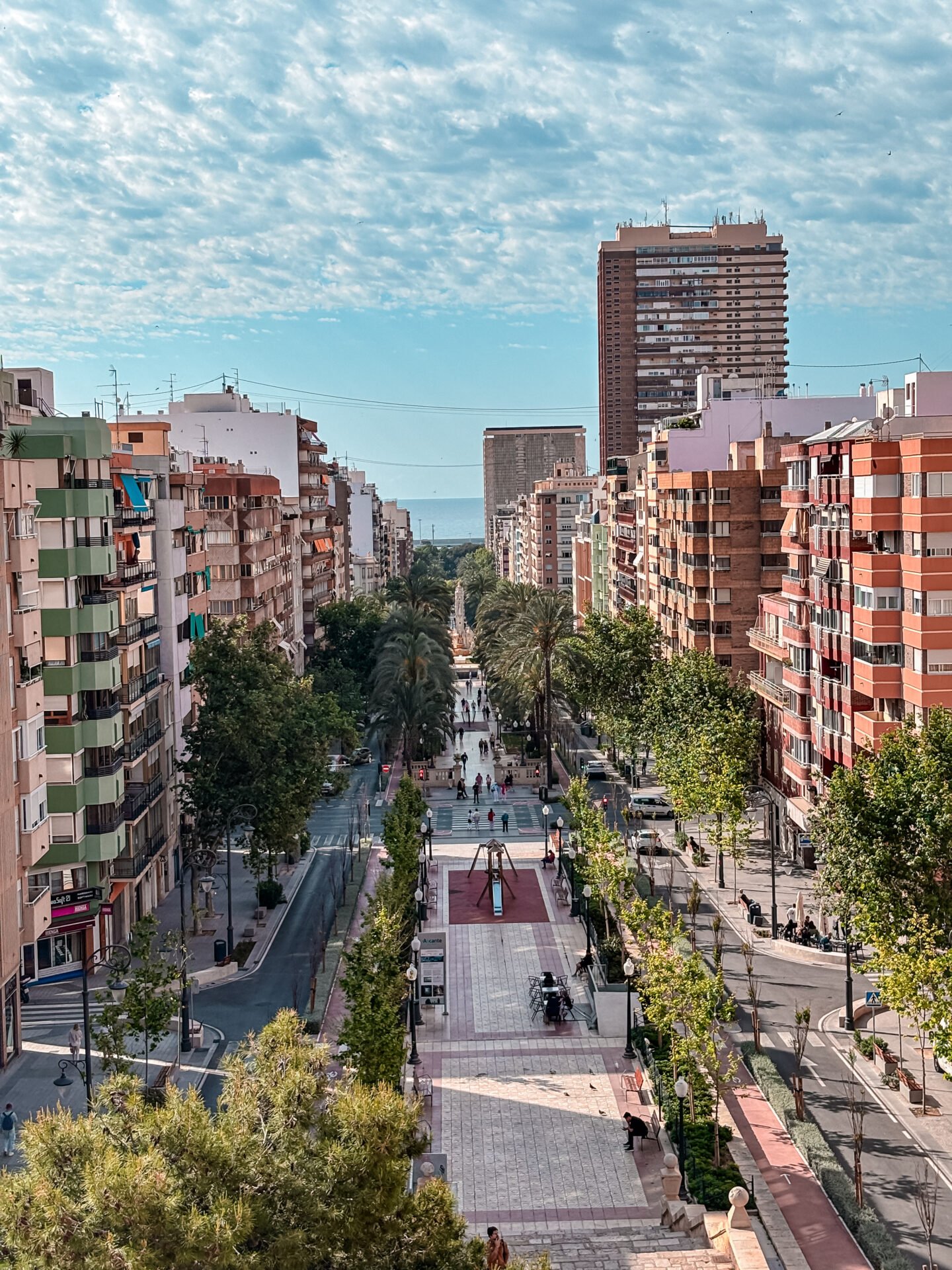 Vista elevada y panor&aacute;mica de la Avenida de la Constituci&oacute;n en Alicante, Espa&ntilde;a. Se muestra un paseo peatonal c&eacute;ntrico flanqueado por &aacute;rboles, edificios residenciales modernos y palmeras. Al fondo, se aprecia el Mar Mediterr&aacute;neo bajo un cielo parcialmente nublado.